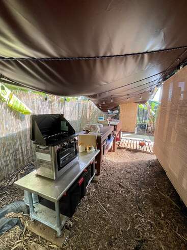 The image depicts a covered outdoor kitchen area with a grill, countertops, and cooking equipment, surrounded by a wooden fence and a chicken coop in the background.