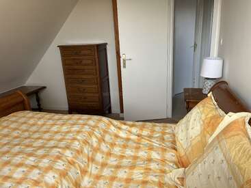 This is a cozy bedroom with a wooden bed, yellow checkered bedspread, tall dresser, bedside table with lamp, and a partially open door leading out.