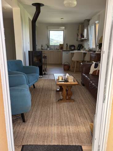 This cozy living space features a blue armchair, brown leather sofa, wood stove, woven rug, coffee table, kitchen area, large window, and countryside view.