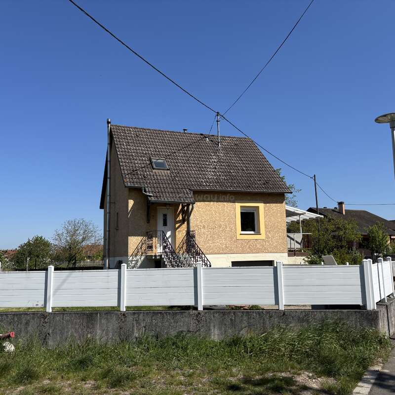 A yellow house with a steep dark roof and white door sits behind a white fence, under a clear blue sky. Trees and grass surround it.