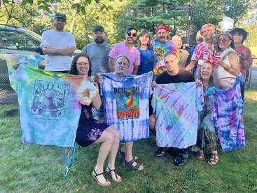 A cheerful group poses outdoors, many smiling and wearing colorful, tie-dye clothing. Several people display hand-decorated shirts, suggesting a fun, creative gathering or reunion.