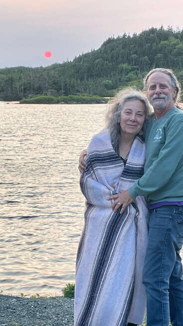 An older couple embraces by a peaceful lake, wrapped in a blanket. The sun sets behind forested hills, casting a serene, romantic atmosphere.