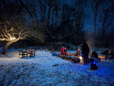 Um grupo de pessoas se junta em volta de uma fogueira numa floresta nevada à noite. Uma luz quente brilha do fogo e de uma lâmpada numa árvore.