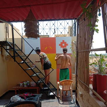 La imagen representa un patio en la azotea con una escalera de caracol, donde dos individuos se dedican a pintar una pared con un diseño geométrico, rodeados de plantas y un toldo rojo.