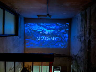 A dimly-lit stairwell with concrete walls features a blue projection that reads "VOICE ACADEMY" over a silky textured background, creating a mysterious and modern atmosphere.