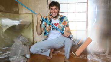 A cheerful man is squatting indoors, holding a paint roller. He wears a floral shirt and smiles widely. The room is covered in plastic, ready for painting.
