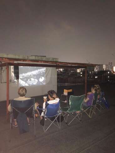 Four people sit in foldable chairs on a rooftop at night, watching a black-and-white movie projected onto a wall, with city lights in the background.