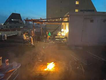 Four people are gathered on a rooftop at dusk, sitting and relaxing near a lit grill, enjoying food, drinks, and each other's company in warm light.