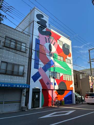 Un grand immeuble est orné d'une fresque murale abstraite aux couleurs vives et aux formes géométriques, avec notamment un motif représentant un œil. Des vélos sont garés à proximité et le ciel est dégagé.