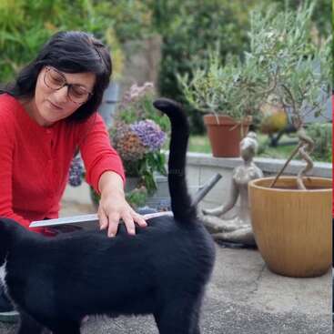 A woman in a red sweater pets a black cat outdoors. She is smiling, surrounded by potted plants and garden decor, enjoying a peaceful moment.