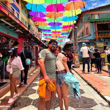 Ein Paar umarmt sich unter farbenfrohen Regenschirmen, die über einer lebhaften, malerischen Straße hängen. Menschen flanieren, Geschäfte säumen den Weg und die sonnige Atmosphäre wirkt fröhlich und festlich.