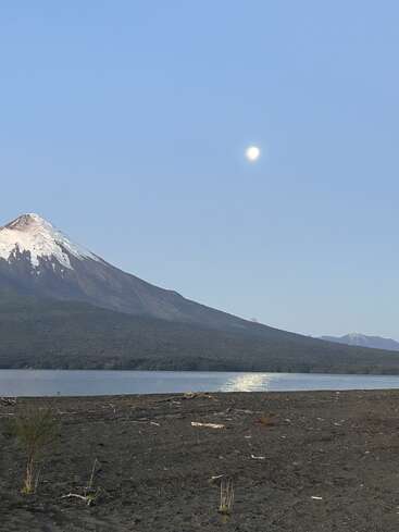 Ein schneebedeckter Berg erhebt sich neben einem ruhigen See. Der Mond scheint von oben und spiegelt sich auf dem Wasser. Im Vordergrund ist ein karges, felsiges Ufer zu sehen. Eine friedliche Szene.