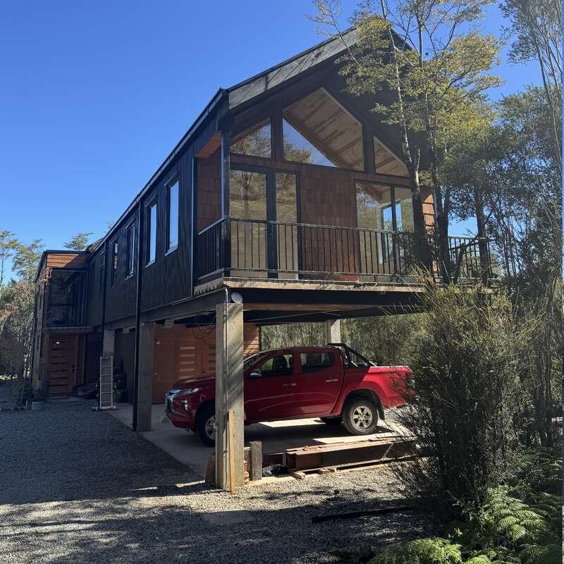 Ein modernes, erhöhtes Haus mit großen Fenstern und Balkon steht zwischen Bäumen. Darunter parkt ein roter Pickup auf einer Schotterauffahrt im hellen Sonnenlicht.