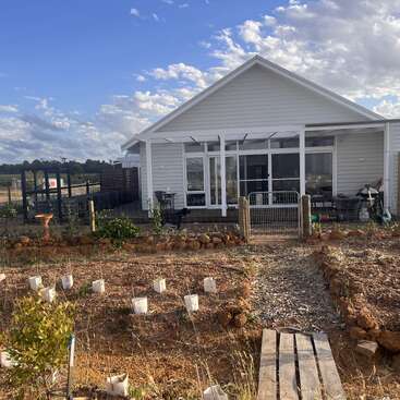 Una casa blanca con porche, rodeada de un jardín vallado con plantas jóvenes y mantillo. El cielo es azul con nubes dispersas en un día soleado.