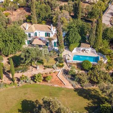 Esta imagem mostra uma villa grande e elegante, cercada por árvores e jardins exuberantes, com piscina, área de pátio e extensos jardins paisagísticos em um ambiente tranquilo.