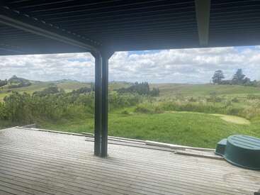 Cette image montre une terrasse en bois sous un patio couvert, donnant sur une campagne pittoresque avec des collines verdoyantes, de l'herbe, des nuages et des arbres éparpillés au loin.
