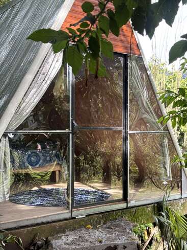 Une cabane confortable à ossature en A, dotée de grandes fenêtres vitrées, reflète la verdure environnante. À l'intérieur, un couvre-lit à motifs et un tapis rond bleu créent une atmosphère paisible et accueillante au milieu de la nature.
