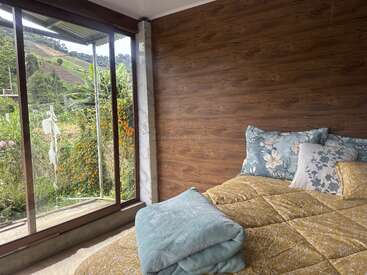 Une chambre confortable avec une literie florale et des murs en bois est dotée de grandes portes vitrées qui s'ouvrent sur un jardin verdoyant avec des collines et des fleurs d'un orange éclatant.