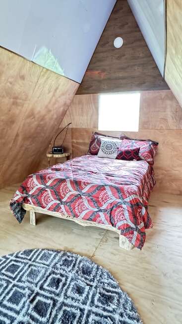 Cette chambre confortable à ossature en A présente un intérieur en bois, un lit double avec des draps à motifs rouges, une petite table d'appoint avec une radio, une fenêtre et un tapis circulaire géométrique gris.
