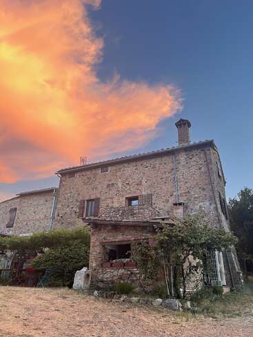 A rustic stone house stands under a dramatic sky with vibrant orange clouds at sunset. Greenery surrounds the building, creating a peaceful countryside atmosphere.