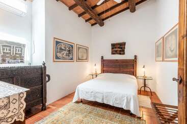 This cozy bedroom features a carved wooden bed, rustic ceiling beams, airy white walls, eclectic artwork, antique furniture, a lacy tablecloth, and plenty of natural light.