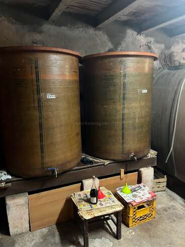 This image shows two large fermentation tanks in a rustic cellar. Two small tables with funnels, bottles, and newspapers are placed in front for bottling.