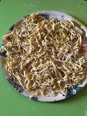 Freshly made, uncooked tagliatelle pasta sits spread out on a round tray, dusted with flour, atop a green table, waiting to be cooked.