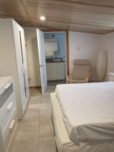 Cette photo montre une chambre bien rangée avec une armoire blanche, une commode, un fauteuil beige, un lit défait, un plafond en bois, un sol carrelé et une porte ouverte donnant sur une salle de bains.