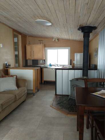Cet espace de vie cosy et compact est doté d'un plafond en bois, d'une petite cuisine, d'un poêle à bois, d'une table à manger, d'un canapé beige et d'un sol carrelé. Ambiance rustique.