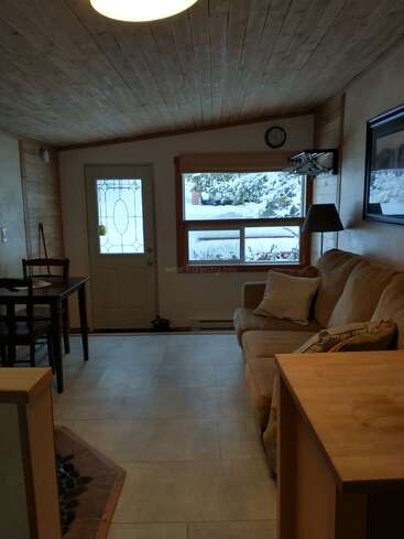 Ce salon confortable est agrémenté d'un canapé beige, d'un plafond en bois, d'une grande fenêtre donnant sur un paysage enneigé, d'une petite table à manger, d'une horloge murale et d'une douce lumière naturelle. Il s'en dégage une atmosphère chaleureuse et accueillante.
