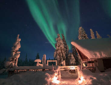 Une cabane d'hiver enneigée brille chaleureusement sous un ciel étoilé, tandis que de magnifiques aurores boréales vertes dansent au-dessus des arbres chargés de neige, créant une atmosphère magique et sereine.