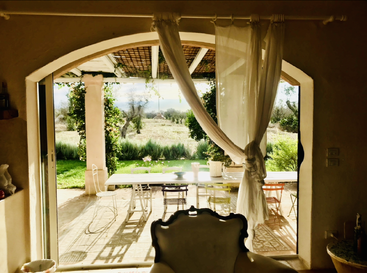 La imagen muestra un sereno patio exterior a través de una ventana arqueada, con una mesa, sillas y una exuberante vegetación, y una cortina blanca que ondea suavemente con la brisa.