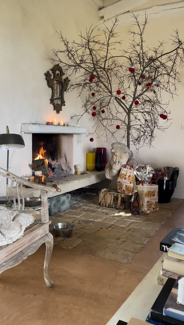 La imagen muestra un acogedor salón con chimenea, un árbol desnudo adornado con ornamentos rojos y una estatua de un hombre, rodeado de regalos y un ambiente cálido.
