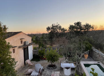 La imagen representa un entorno sereno al aire libre, con una casa, una piscina y un patio, rodeados de árboles y un muro de piedra, con una pintoresca puesta de sol al fondo.