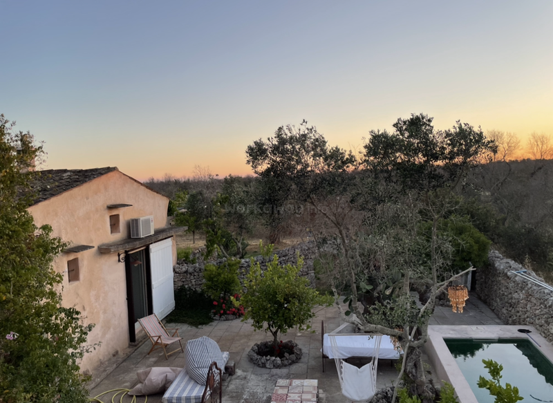 La imagen representa un entorno sereno al aire libre, con una casa, una piscina y un patio, rodeados de árboles y un muro de piedra, con una pintoresca puesta de sol al fondo.