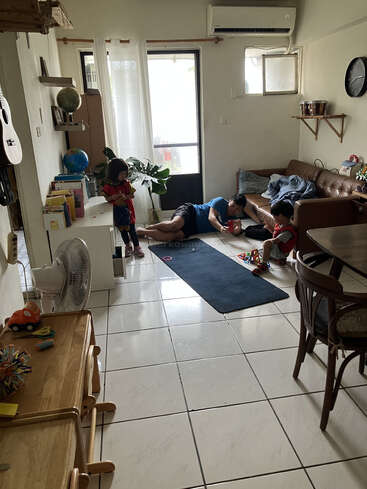 Un salon confortable avec un homme allongé sur le sol, deux enfants qui jouent, des étagères, un canapé, des plantes, des jouets et la lumière naturelle de la fenêtre.