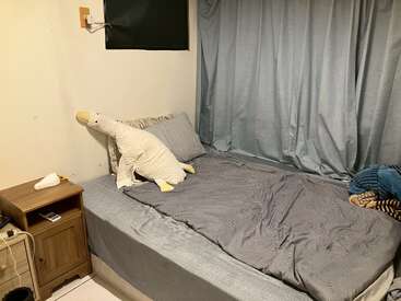 Un lit bien fait avec une literie grise, un canard en peluche sur les oreillers, des rideaux gris, une table de nuit en bois avec la télécommande, une boîte à mouchoirs, et une atmosphère douillette.