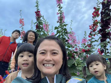 Uma família posa para uma foto em frente a altas flores desabrochando sob um céu azul. Todos estão sorrindo e apreciando juntos a bela paisagem ao ar livre.