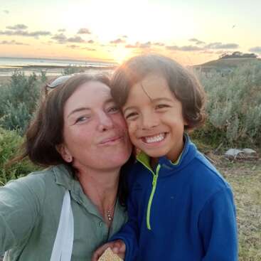 Uma mulher e uma criança sorriem alegremente para uma selfie ao ar livre, com uma vegetação exuberante e um belo pôr do sol na água ao fundo. Momento de alegria.