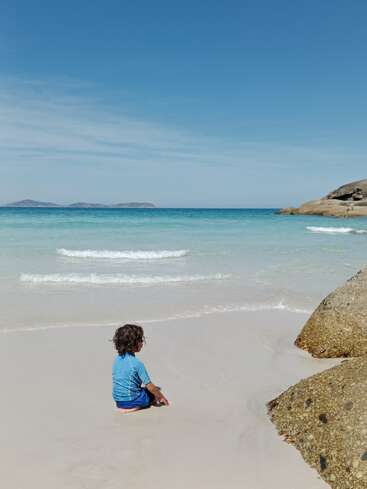 Un niño pequeño sentado en la suave arena blanca de la playa, contemplando el tranquilo mar turquesa, bajo un cielo azul claro con islas lejanas.