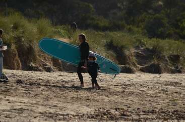 Una mujer y un niño caminan por una playa de arena, llevando una tabla de surf azul. Ambos llevan trajes de neopreno. Al fondo, arbustos y árboles verdes. Día soleado.