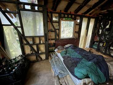 Dieses Bild zeigt eine rustikale Hütteneinrichtung mit einem ungemachten Bett, Holzregalen, Flaschen und mattierten Fenstern. Natürliches Licht fällt ein und verleiht dem Raum eine gemütliche Atmosphäre.