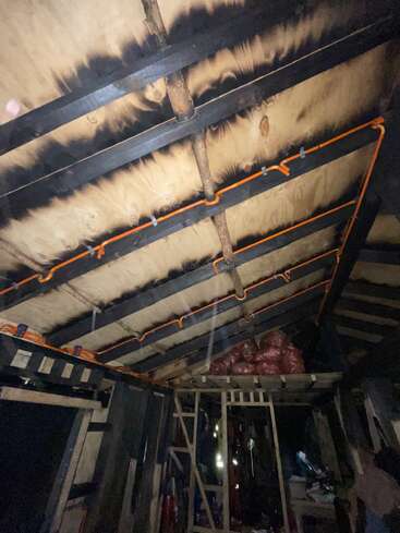 Dieses Bild zeigt das Innendach einer Holzkonstruktion mit sichtbaren schwarzen Brandspuren, orangefarbenen Drähten, Balken und auf einem Dachboden gelagerten Taschen.