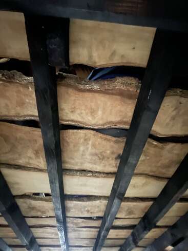 Dieses Bild zeigt eine Nahaufnahme von rauen Holzbrettern mit natürlichen Kanten, die von schwarzen Balken gestützt werden, wahrscheinlich von der Unterseite eines rustikalen Tisches oder einer Bank aus gesehen.