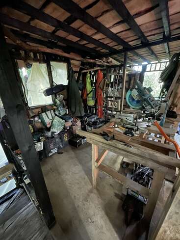 Dieses Bild zeigt eine unübersichtliche Werkstatt mit hölzernen Werkbänken, Werkzeugen, Schutzausrüstung, Kisten, einer Kreissäge, Schutzkleidung und verstreuten Materialien in einer rustikalen, teilweise offenen Struktur.