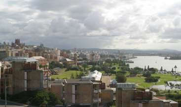 A imagem mostra uma paisagem urbana com um parque, um corpo de água e um céu nublado. A paisagem urbana apresenta uma mistura de edifícios e árvores.