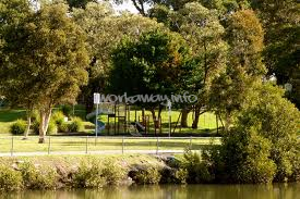 A imagem mostra uma cena serena de um parque, com um playground, árvores e um corpo d'água, com uma sutil sobreposição de marca d'água.