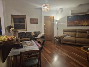 A cozy living room with wooden floors, sofas, colorful artwork, a small dining table, floor lamp, fruit basket, and a child sitting comfortably on the couch.