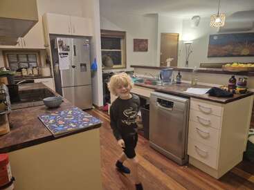 A cheerful blond child walks energetically through a cozy, well-lit kitchen with modern appliances, wooden floors, colorful artwork, and various utensils and dishes visible.