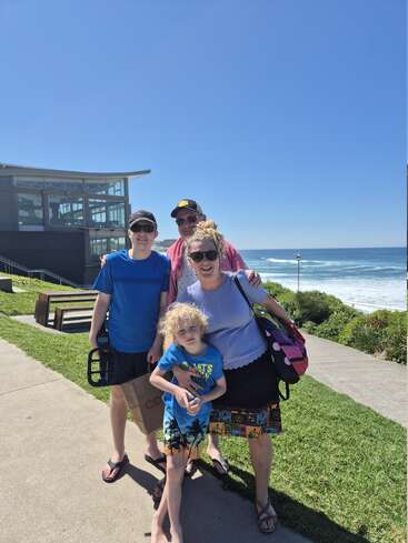 A family of four poses happily on a sunny day near the beach, dressed for summer, with ocean waves and a modern building in the background.
